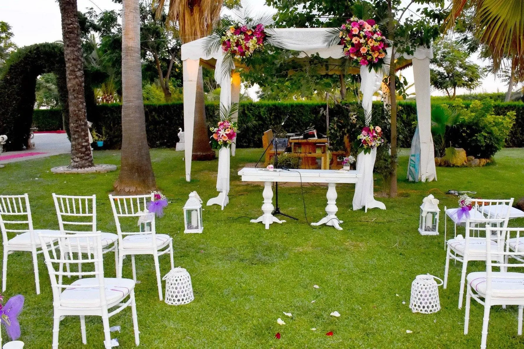 Un altar blanco de tul y flores sobre el césped, con sillas blancas delante y una mesa de madera