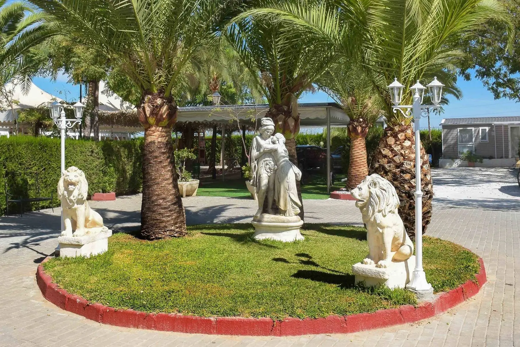 Una glorieta de césped con estatuas de piedra blanca, dos leones y una farola de tres globos entre palmeras