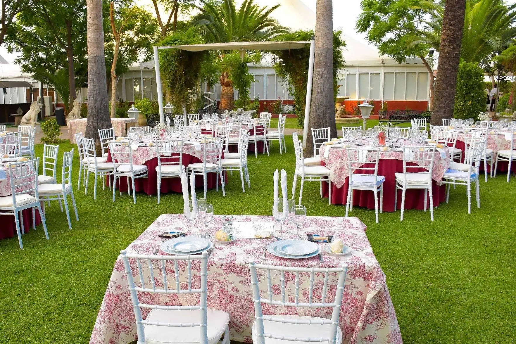 Mesas de banquete con manteles rosas sobre el césped, con una pérgola blanca y la carpa al fondo
