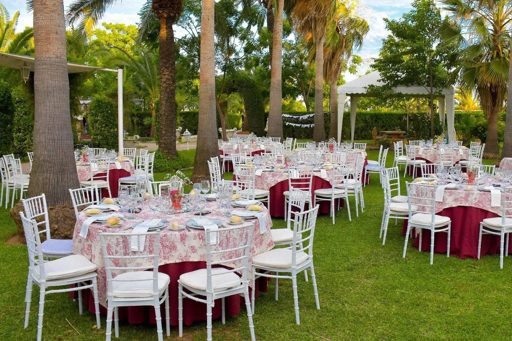 Mesas redondas con mantel de toile rosa y sillas blancas sobre el césped, bajo palmeras