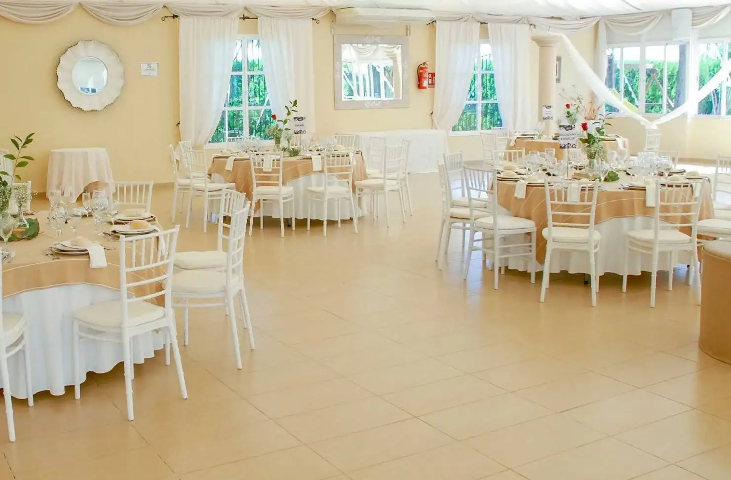 El salón de banquetes con ventanales al jardín, mesas redondas con manteles color arena y sillas blancas