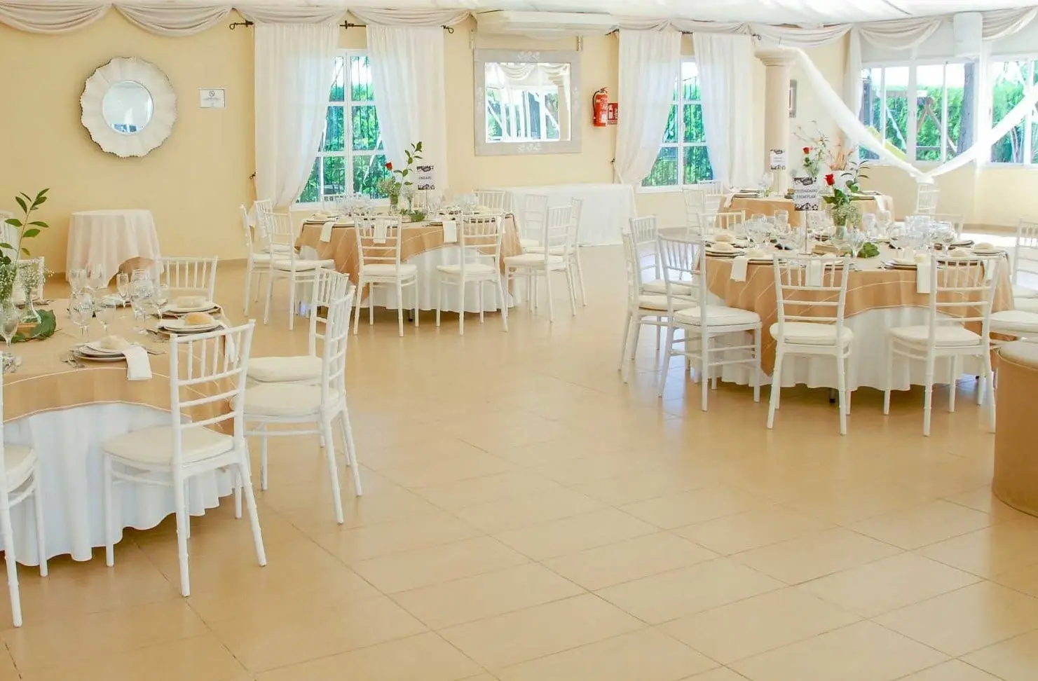 El salón de banquetes con ventanales al jardín, mesas redondas con manteles color arena y sillas blancas